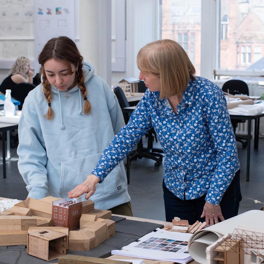 A student and a tutor are standing behind a table looking at cardboard model of a building in front of them. The tutor is gesturing towards the model.