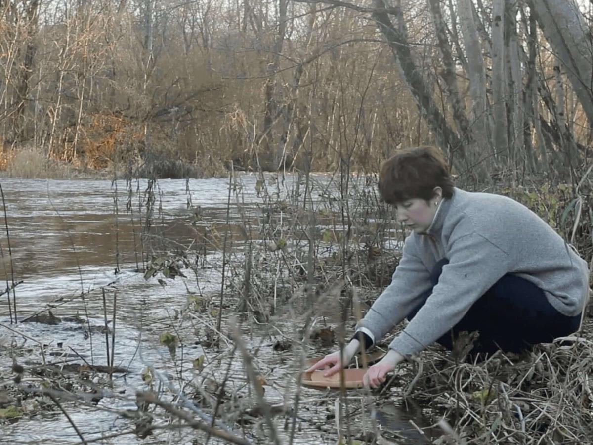  Digital Film Still of Installation, a person crouches by the water edge holding a piece of sculpture in the water.