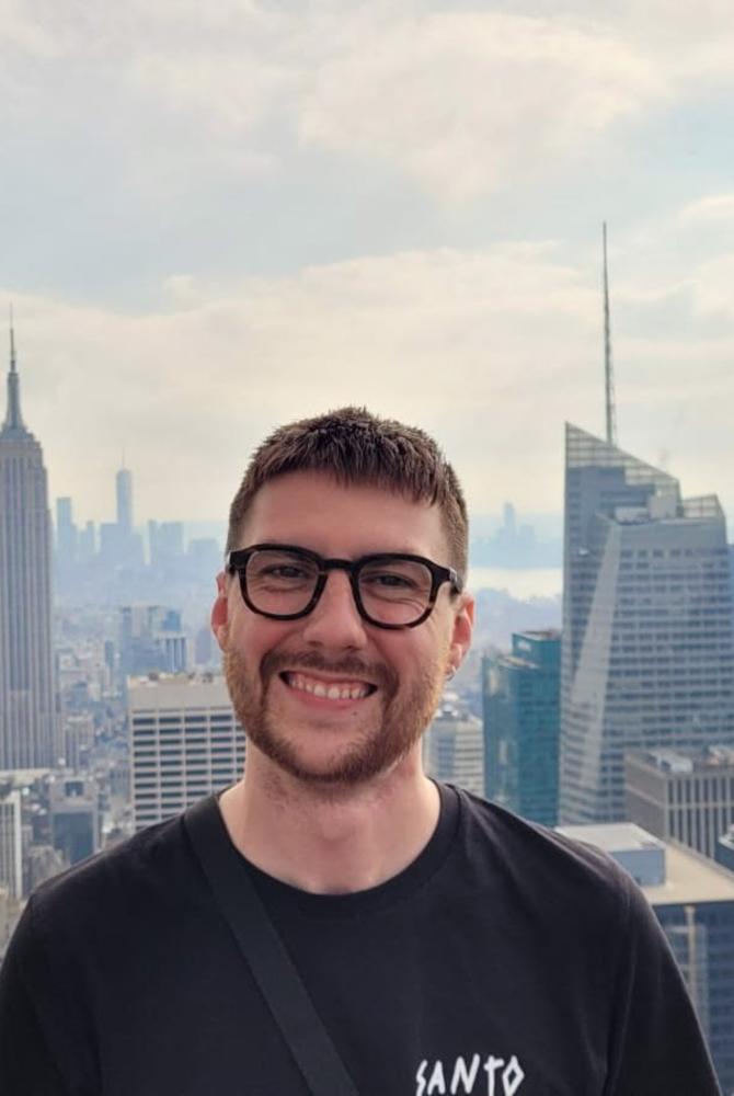 A person wearing a black t-shirt and glasses, with the New York skyline behind them.