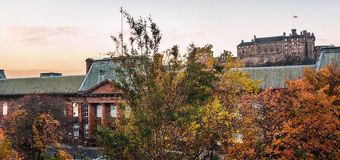 The Edinburgh College of Art Main Building during the autumn with trees in the foreground and Edinburgh Castle in the background
