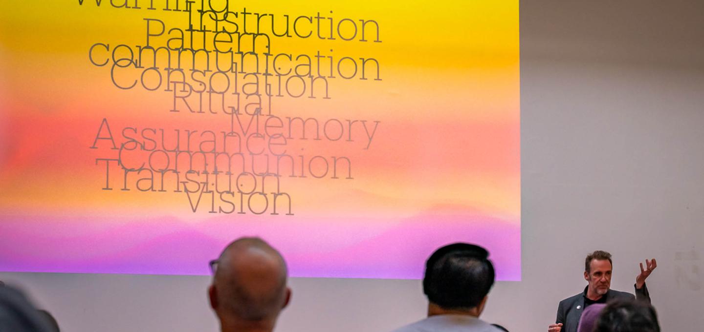 An event speaker gesturing to the side of a colourful projected slide in yellows, pinks and violet.