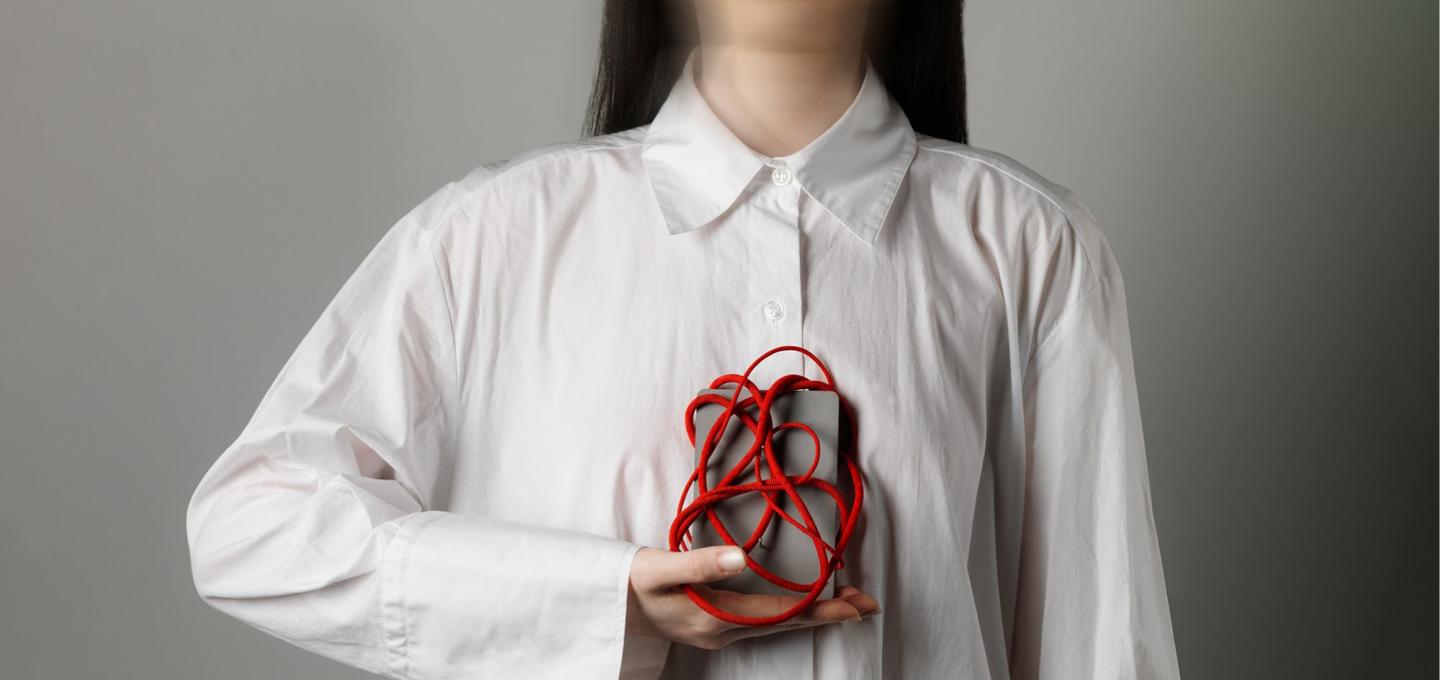 Person wearing a white shirt holds object to chest. The object is a grey box wrapped in a tangle of red cord. 