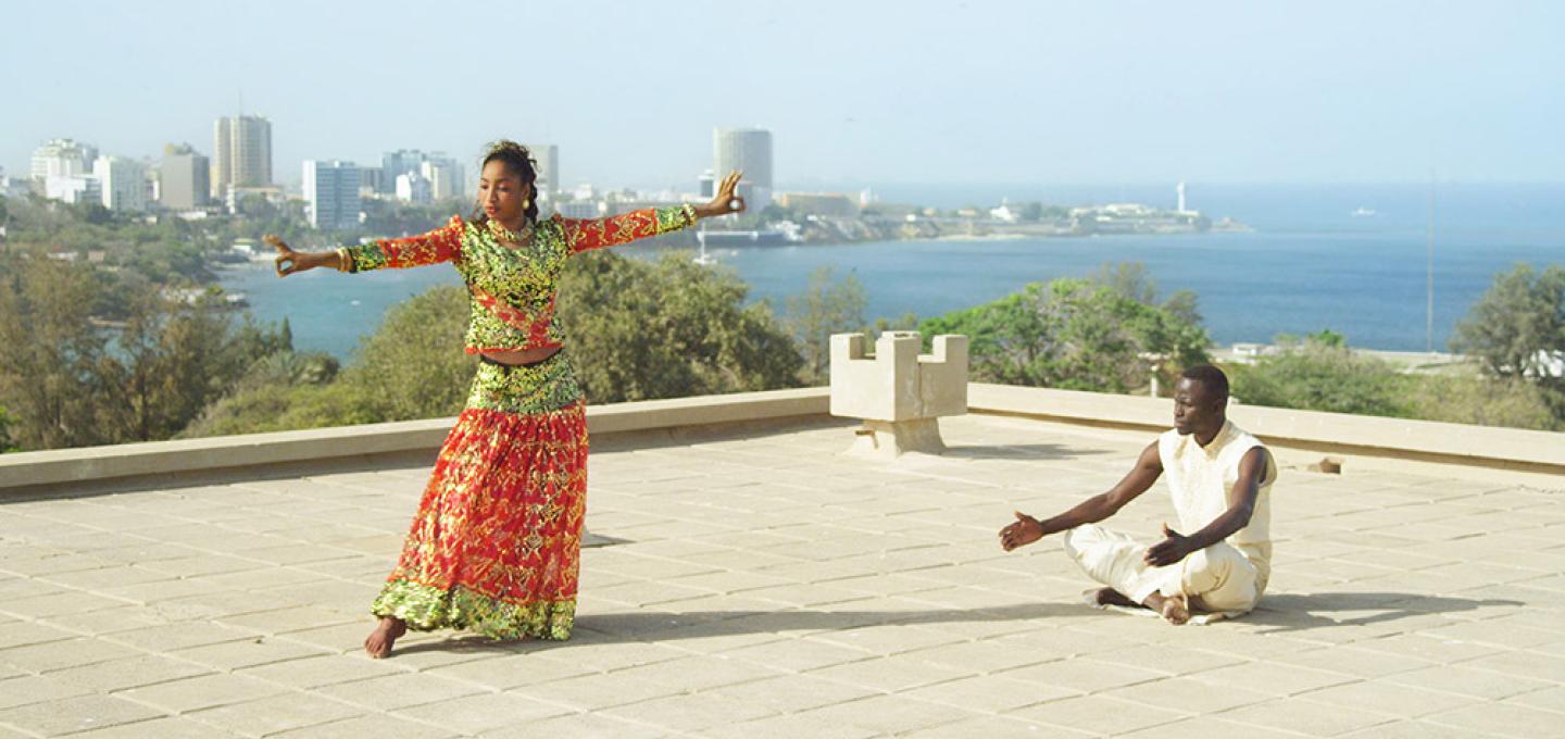 A black woman dancing in colourful clothing and a black man in white, sitting cross-legged, stand on a large rooftop with a modern cityscape by the sea as a background.
