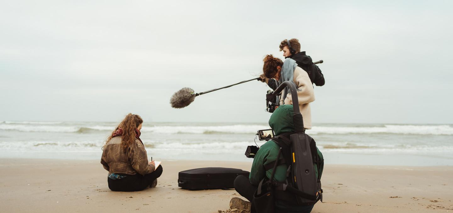 Camera crew with sound recordist filming a woman sitting on the beach, writing.
