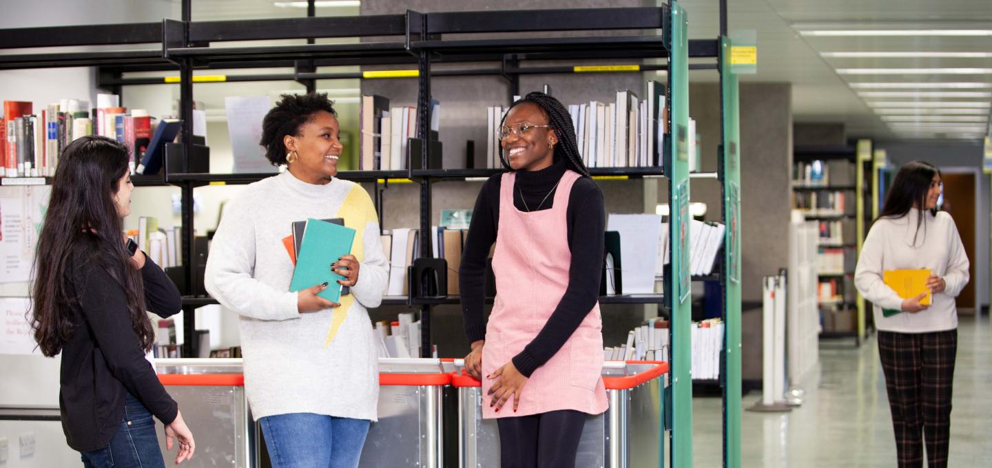 Three female students chatting and smiling in a library, with another female student walking in the background