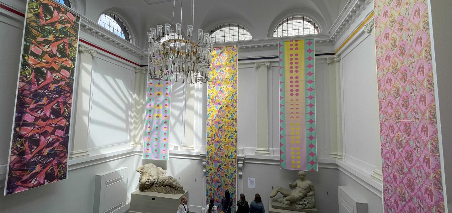 The staircase of Edinburgh College of Art's Main Building, with five pieces of textiles work hanging from the high ceilings and sunlight coming through the skylight. 