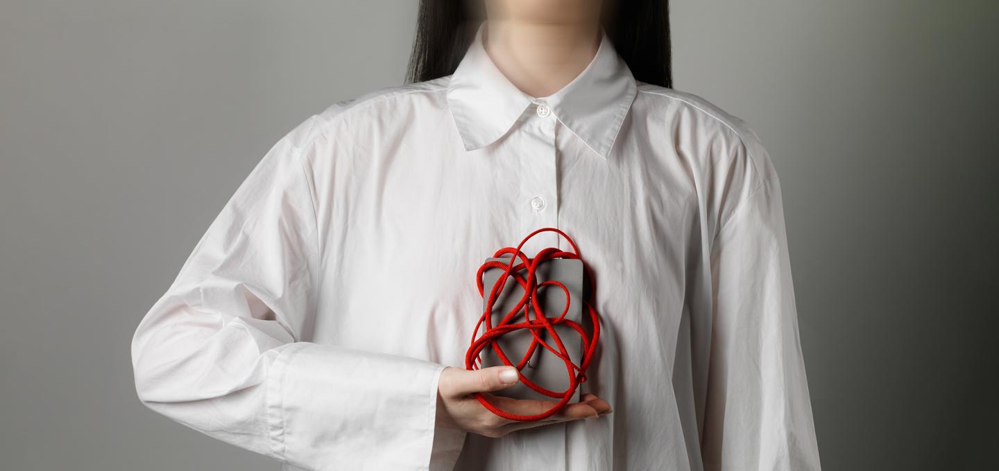 Person wearing a white shirt holds object to chest. The object is a grey box wrapped in a tangle of red cord. 