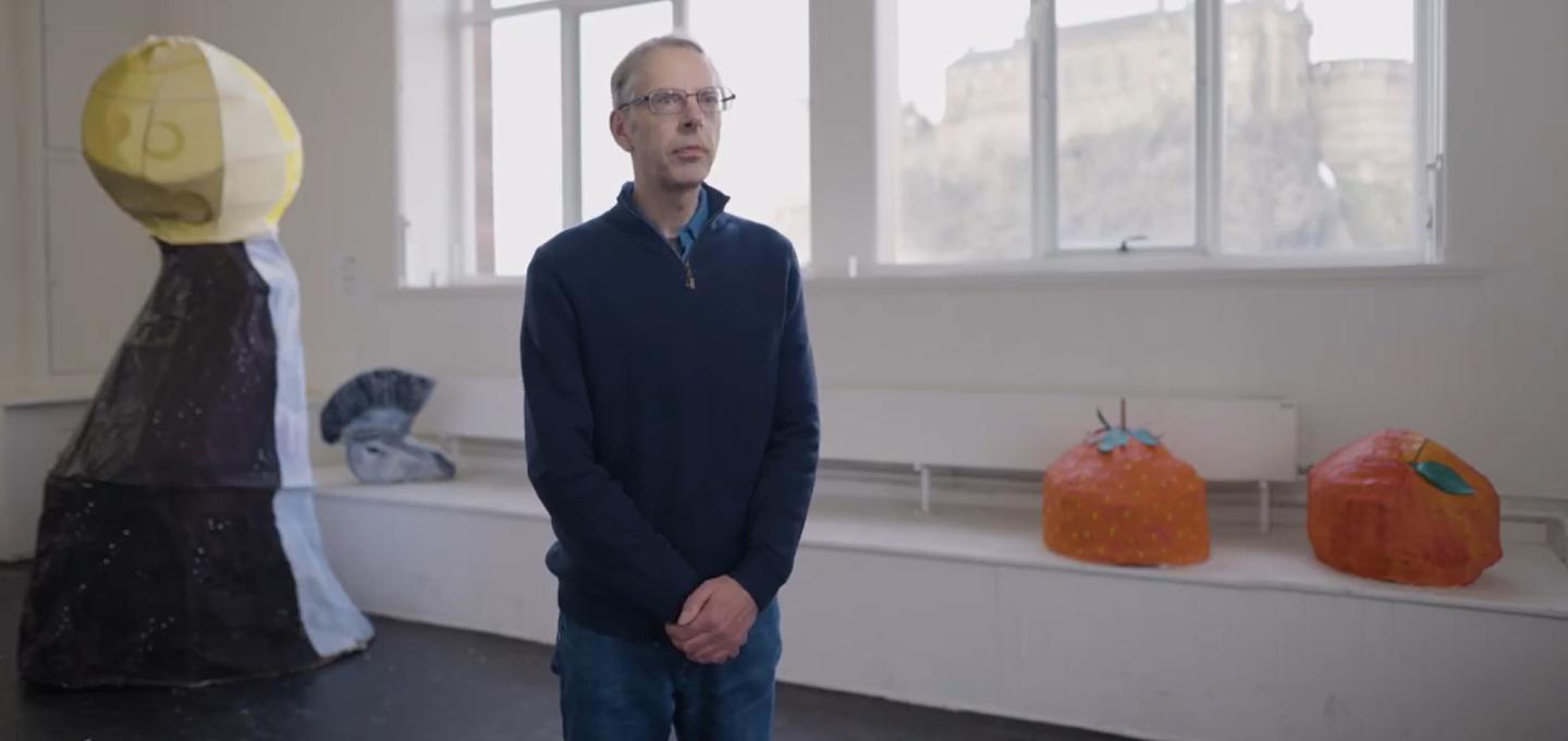 Lecturer of Fine Art, John Brown, stands in the middle of one of the ECA painting studios. He has a few pieces of art behind him including two pumpkin-like objects and a tall, curved object with a yellow ball atop.