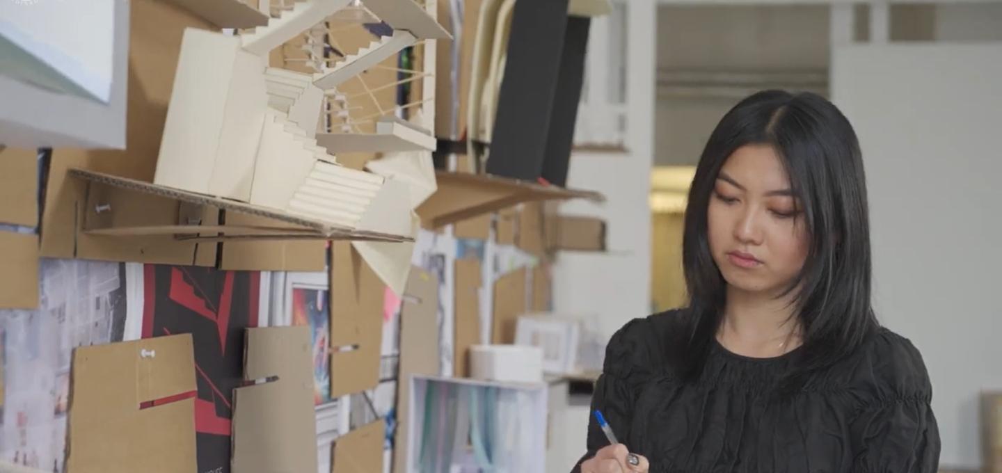 A student writes something down as they stand by a wall with multiple models of interior spaces.