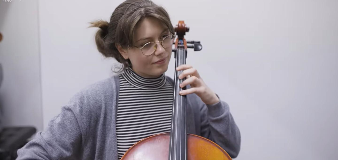 A student on the Musicology programme plays the cello. They are wearing a striped polo neck top and a grey cardigan.
