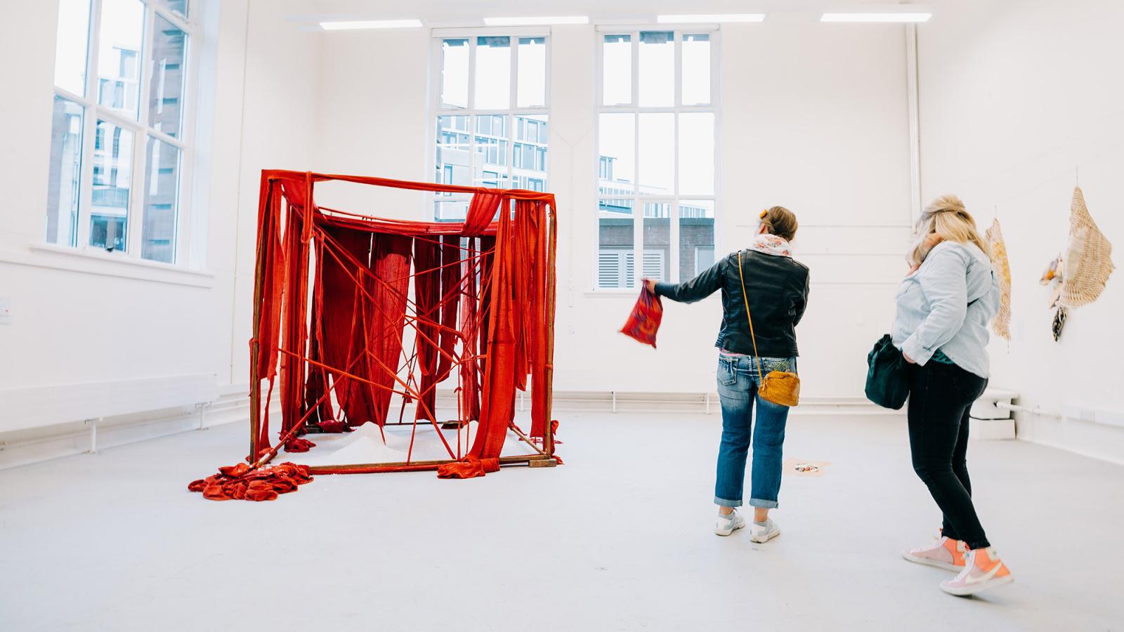 Two visitors to the June 2023 Graduate Show are looking at a large red textile installation in a white studio with large windows.