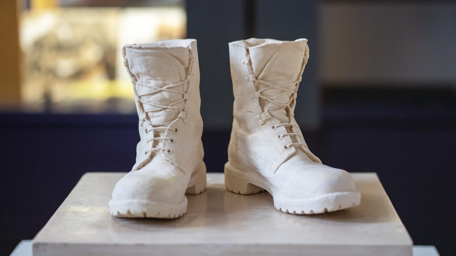 A cast of a pair of combat boots. They're unlaced and slightly worn; an off-white colour standing on a marble pedestal
