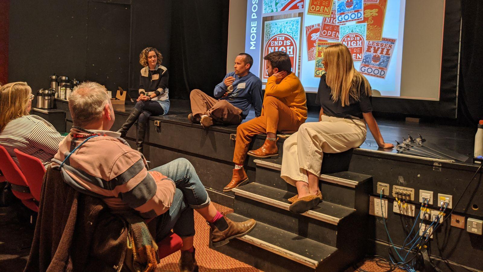 Four people sitting on the edge of a stage in discussion, one holding a microphone. Audience members in front of them are listening; behind them is a projected backdrop of colourful illustrations