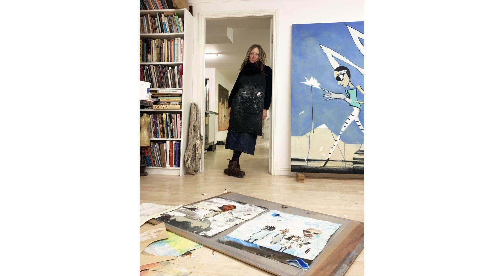 Moyna is standing in the doorway to a her studio, leaning on the door frame. She is dressed in black, with white paint marks on the front of her garments. To one side of her is a full bookshelf, to the other a striking, large blue painting of a woman in front of what looks like a white, electrical flash in the sky. Two smaller pieces of work are on a drawing board in the foreground, each featuring painted body parts.
