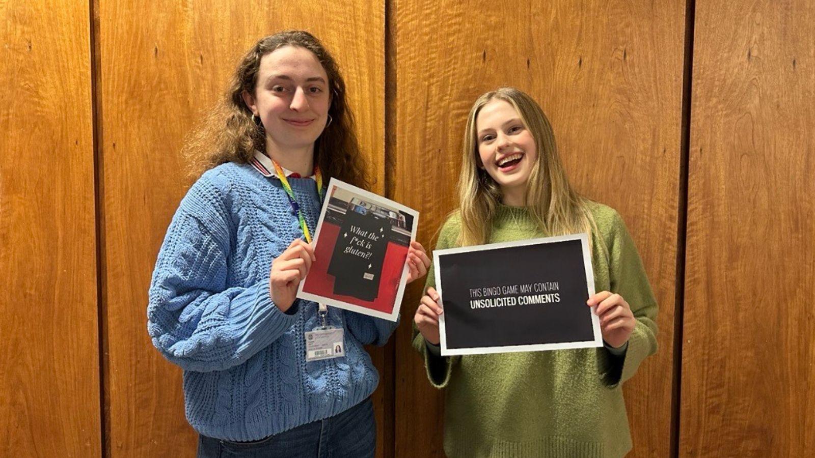 Chiara and Katie standing side by side, smiling at the camera. They are each holding a piece of work from their competition entry. Chiara holds a photo of a tea towel with the words 'What the f*ck is gluten?' on it, Katie holds a black sheet with white lettering, reading 'This bingo game may contain unsolicited comments'.