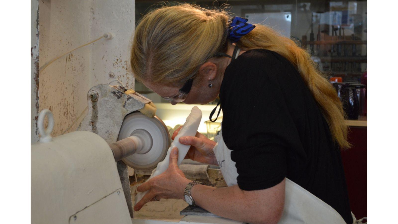 A woman is polishing a white, sculpted piece of glass on a spinning polishing machine. She is wearing safety glasses, and looking closely at her work.