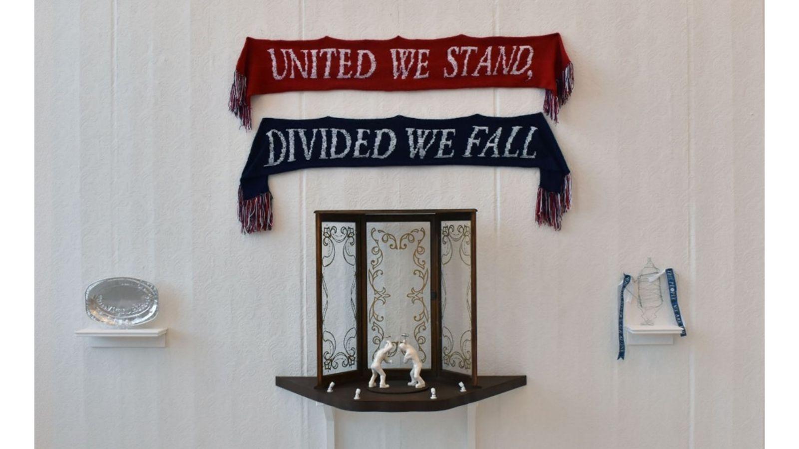 A white gallery wall. At the top are pinned two scarves with white text on. The top one is red and reads 'United we stand,' the bottom one is blue and reads 'Divided we fall'. Beneath is a shelf holding a 3D frame and two white figures. There are smaller shelves either side, one holding a silver-coloured platter, the other a trophy with blue ribbons from the handles.