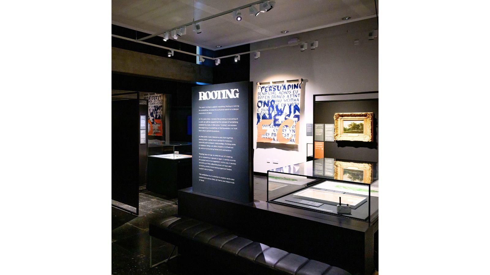 Interior shot of the Rooting exhibition - a black panel bears white text, headed Rooting, there are paintings and display cases behind.