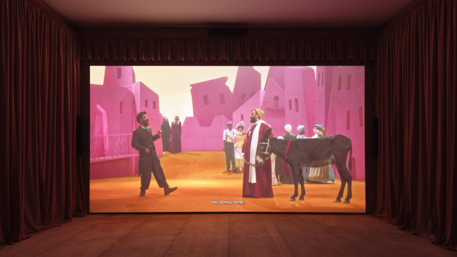 A scene from a film by artist Wael Shawky. Two people are talking to each other. One of them holds the reigns of a donkey. Other people stand nearby and they are surrounded by pink stone buildings.