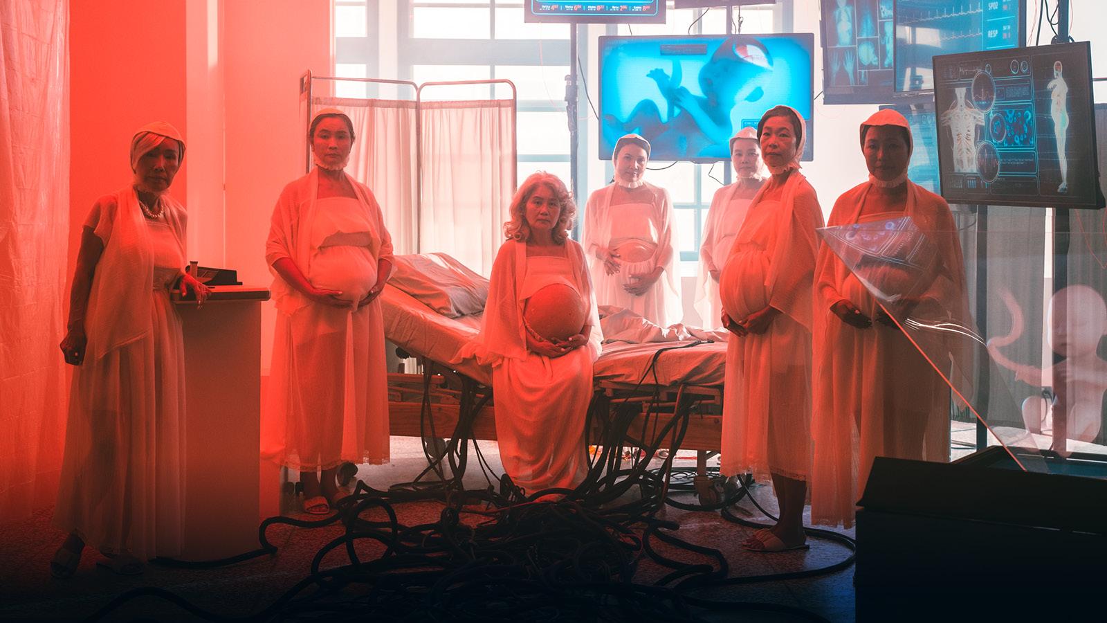 A photo of 7 pregnant people standing in white robes, in a room set up like an operating theatre with a hospital bed, hospital screens and large plasma tv screens with diagrams and data on them.