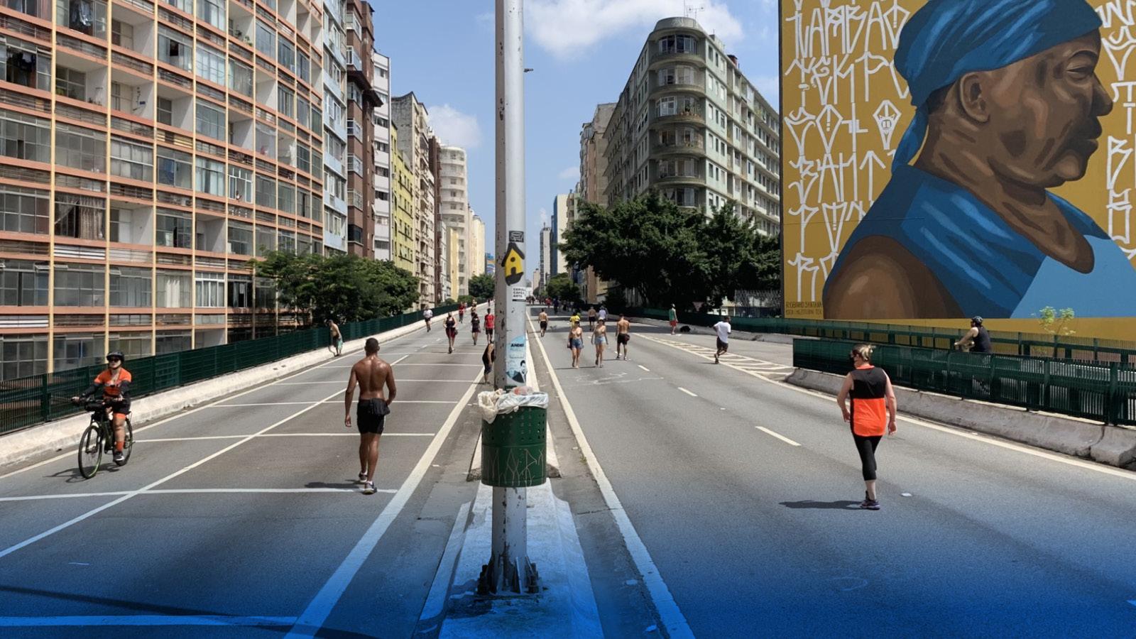 An expressway surrounded by tall buildings stretches into the distance. People walk in the middle of the road. One building on the right has a large mural of a person wearing a blue sleeveless top, a blue wrap around their head, where a green container balances.