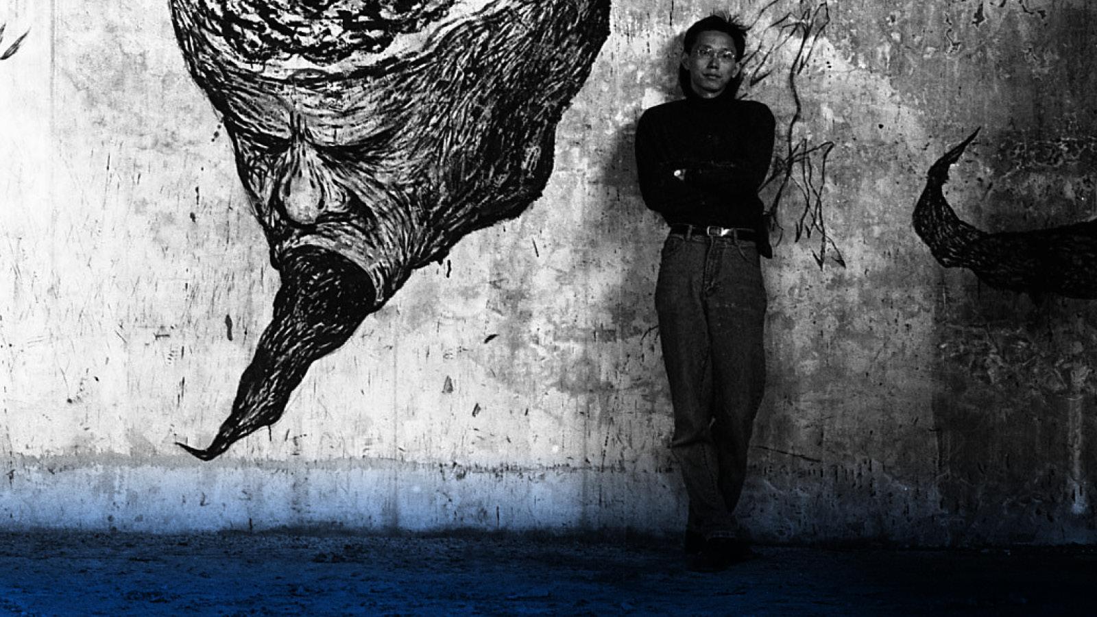 A black and white photo of a person leaning against a wall next to a mural of a face with a black mass pouring from its mouth.