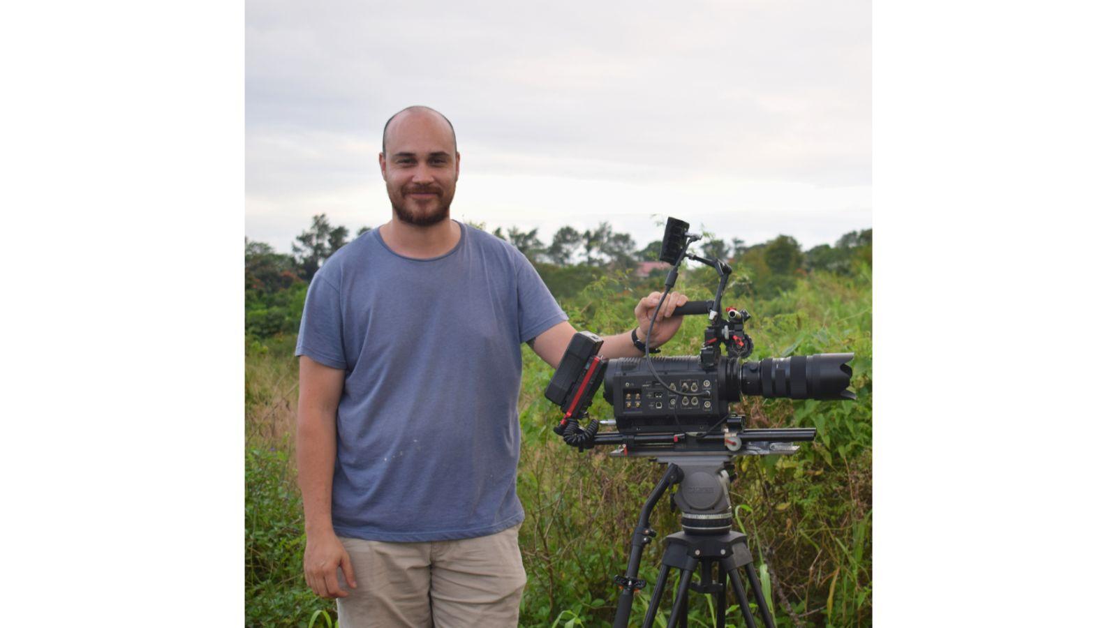 Andrew stands beside the camera, in a field of lush greenery