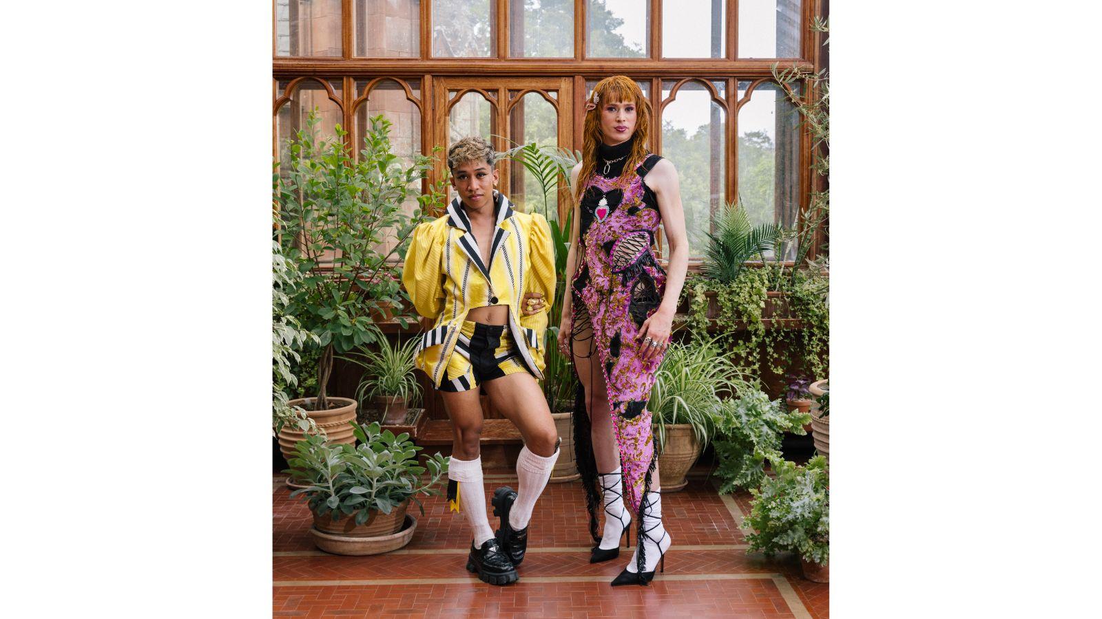 Davide and Angel stand in front of a wall of windows, lush green plants either side of them. Davide wears an elaborate yellow jacket and shorts, Angel a pink and black dress with black lacing.