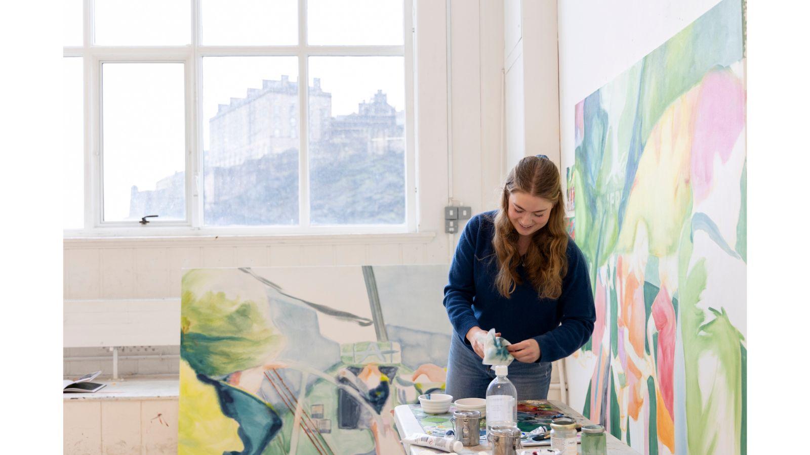 Phoebe in her studio at ECA. A colourful painting is on the wall beside her, another on the floor behind her. She looks to be cleaning brushes. The Castle is visible through the window behind her.