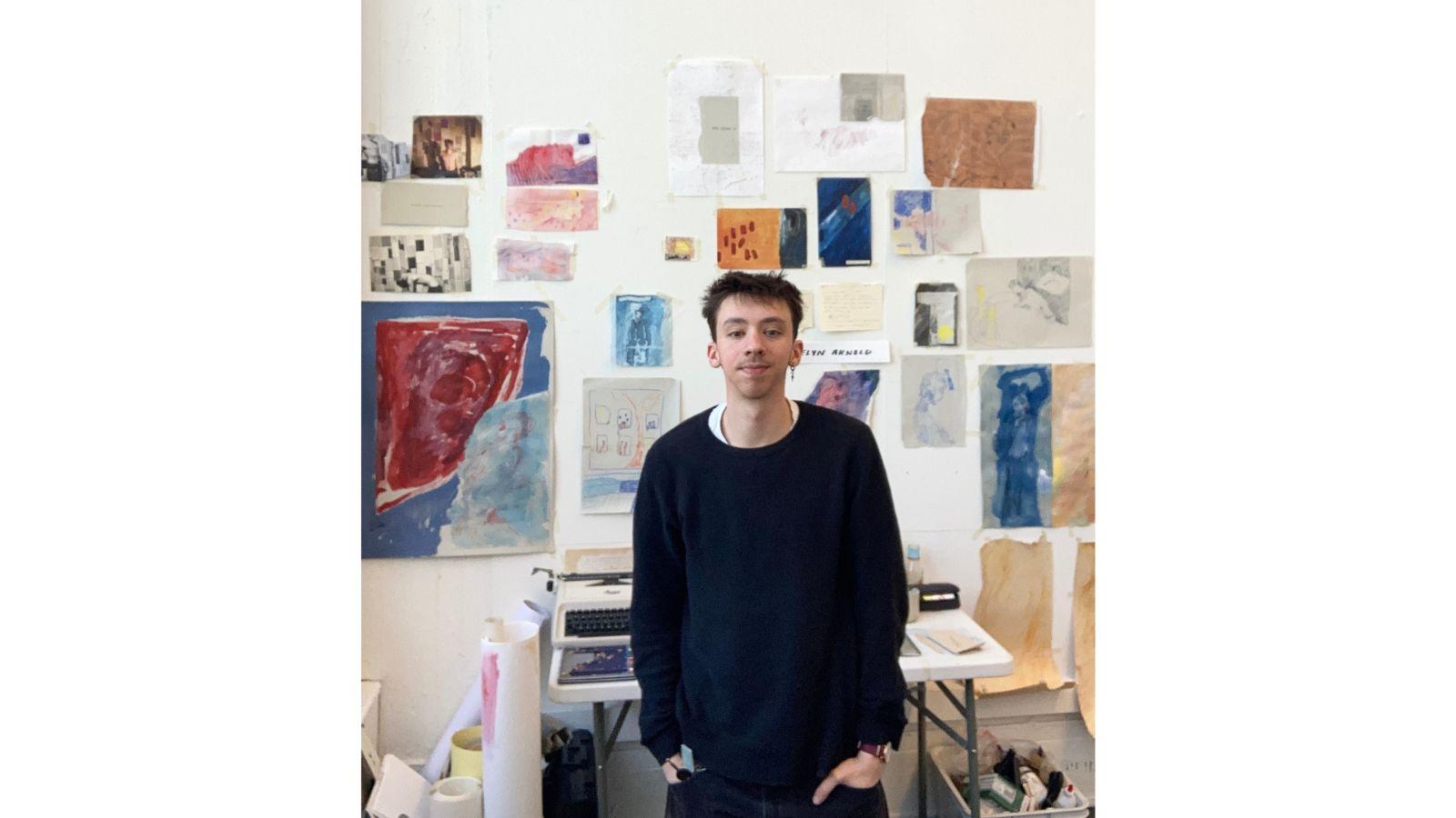 Thomas, wearing a black jumper, stands in his studio, looking at the camera. The white walls are adorned with paintings on sheets of various sizes. Behind him is a table, with a typewriter on it.
