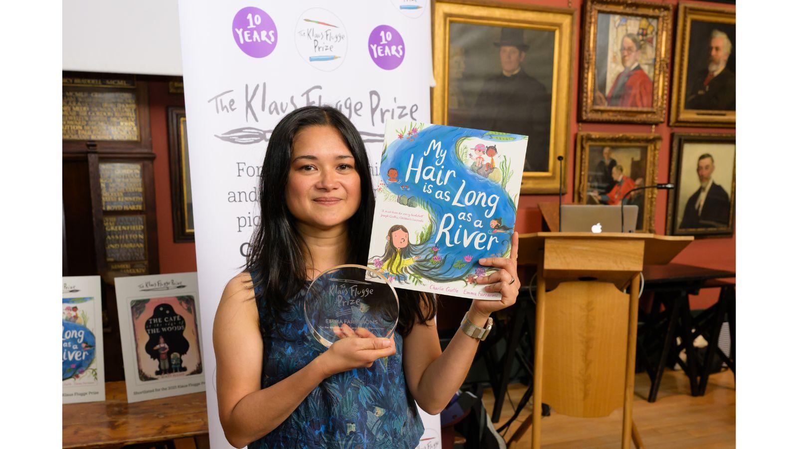 Emma holds a copy of the book, and her glass award. On the wall behind her are several framed portraits.