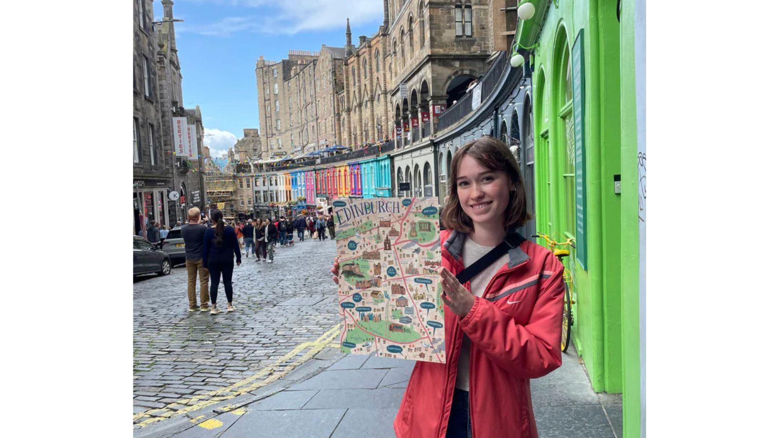 Ally stands in front of the coloured shop fronts of Victoria Street in Edinburgh, holding up one of her illustrated maps