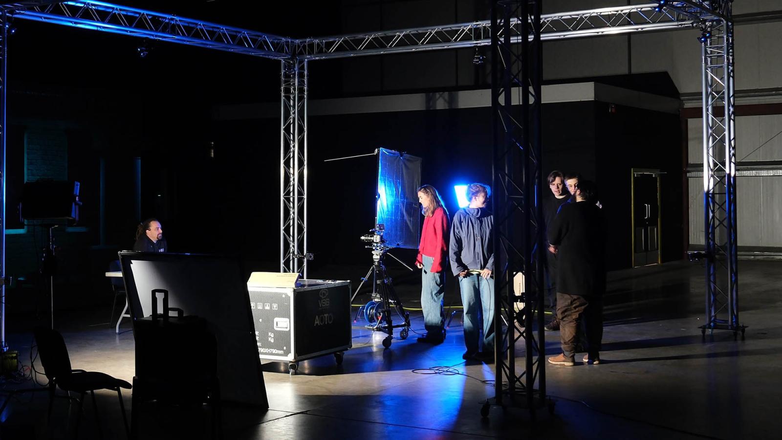 A small group of students are working with camera equipment. The studio is mainly dark but there are some dramatic spotlights.