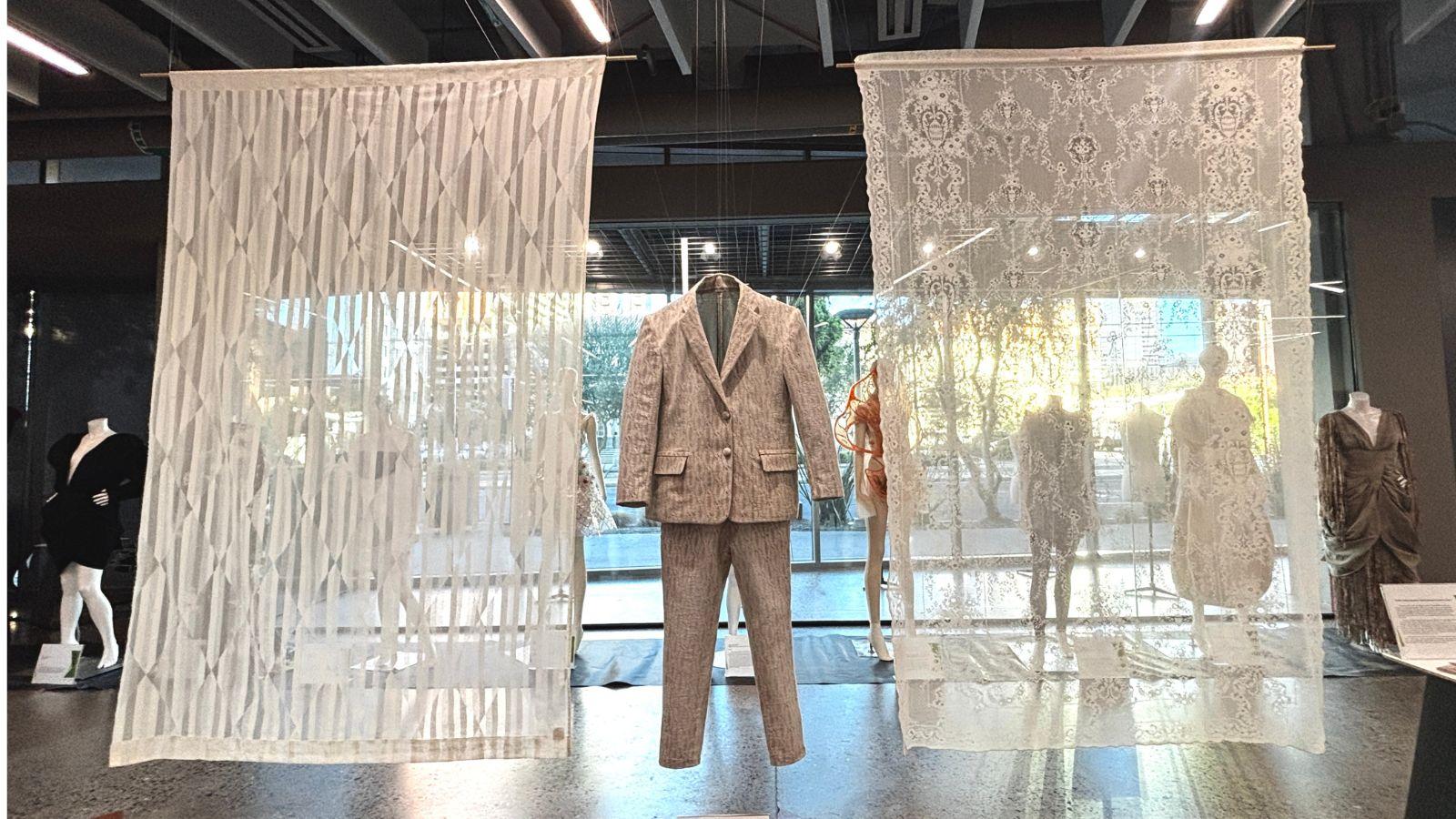 The suit, made of lace, hangs from the ceiling of an exhibition space. Either side of it are sheets of lace, the one on the left with a pattern of lines and diamonds, the one on the right much more traditional.