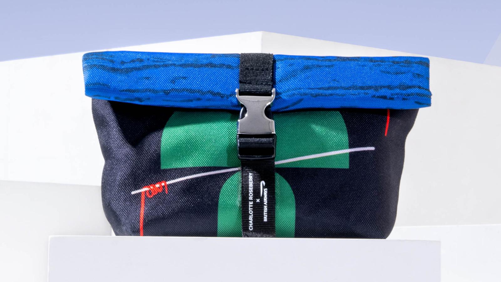 A fold-over bag with green graphic shapes on a black background and blue top. It is secured by a black strap and silver modern buckle.