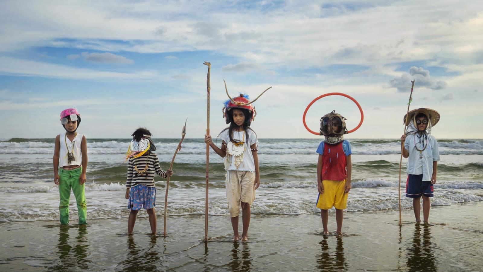 Five children standing at the shoreline. They hold sticks and wear and wear various styles of head gear.