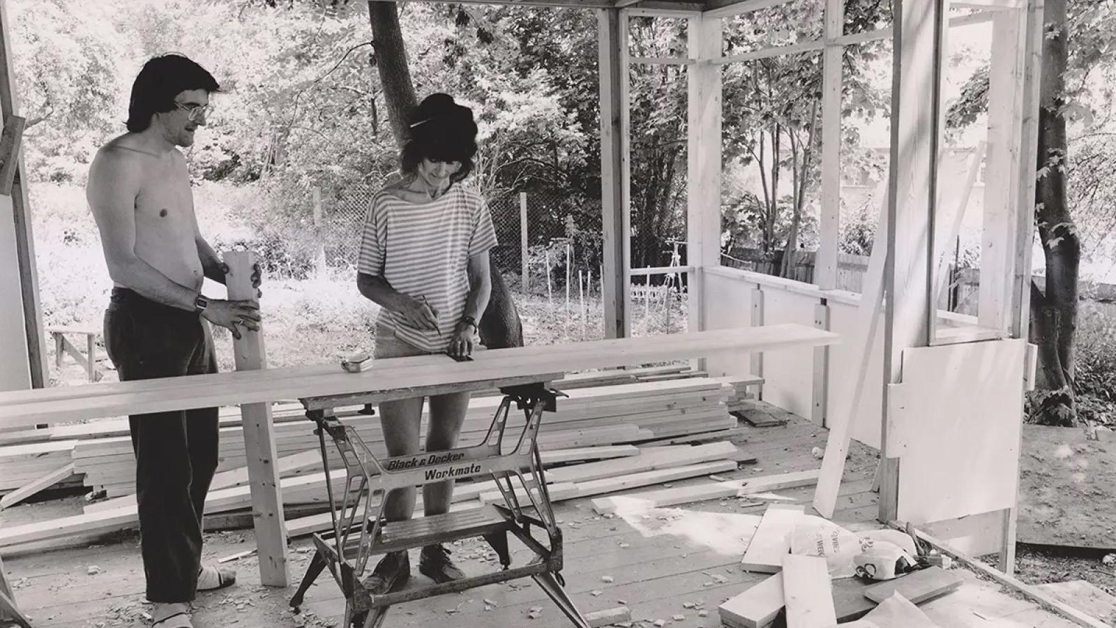 A black and white photo of two people on a building site of a house. One is sawing wood, the other watches. 