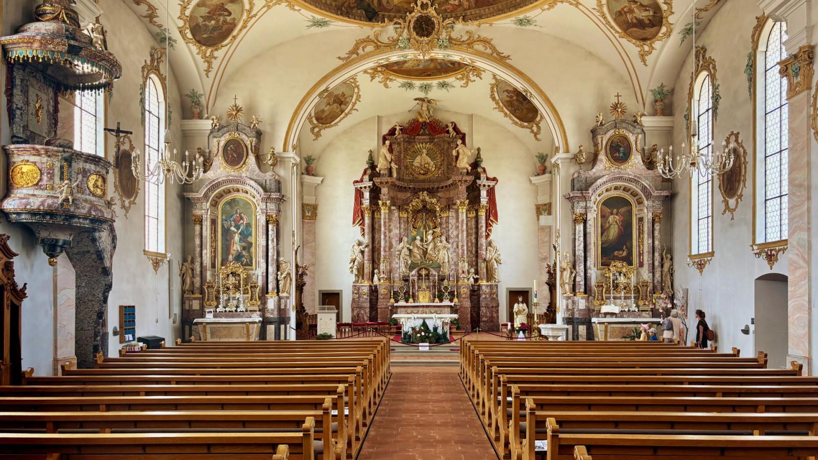 An image of the interior of the Heiligkreuzkirche in Kirchberg taken from the back of the church looking towards the altar. There are rows of wooden pews under the domed ceiling that's adorned with a fresco and gold, ornate cornicing and decoration.