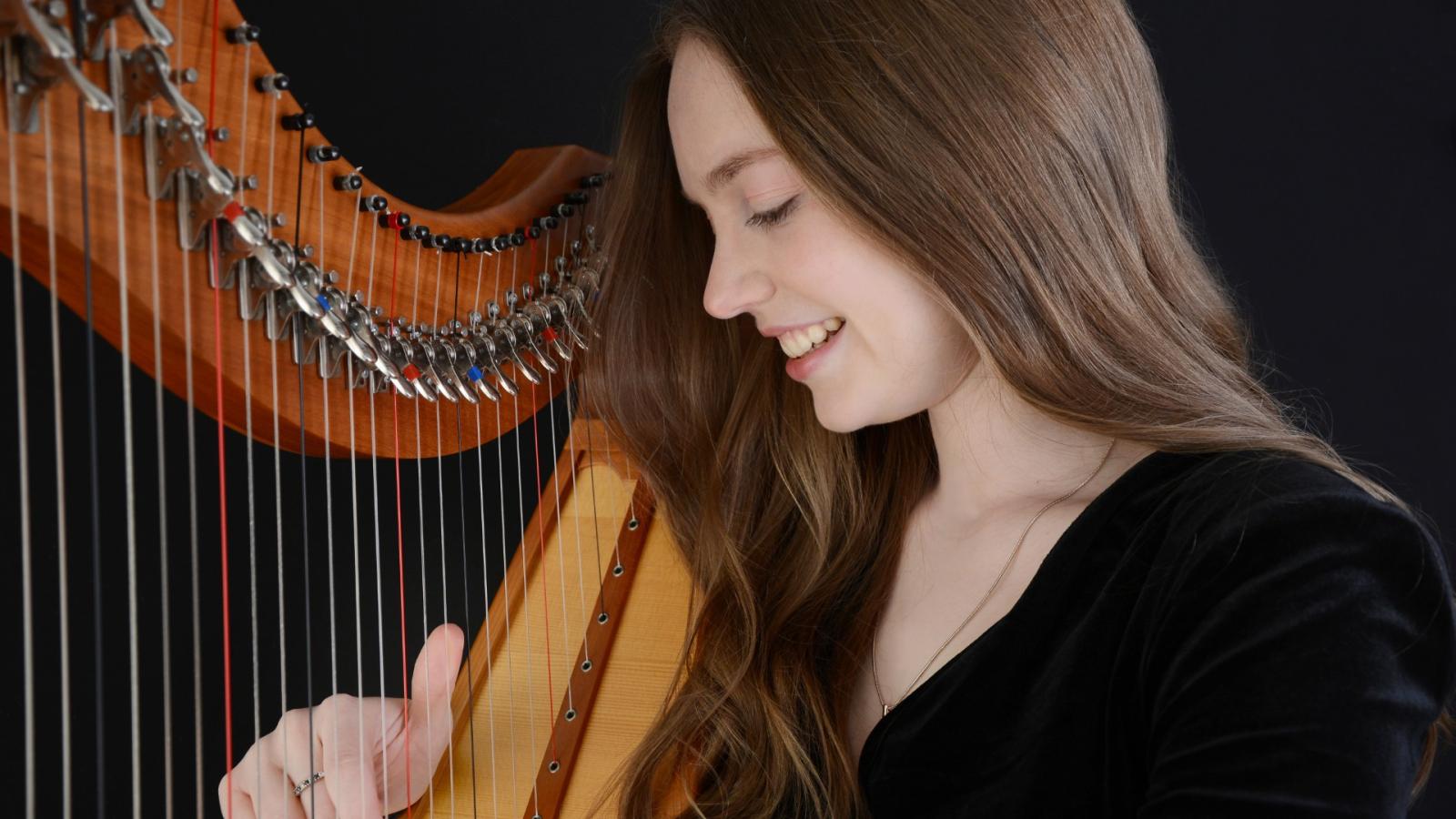 A person with long brown hair is looking down and smiling. They are playing a harp.