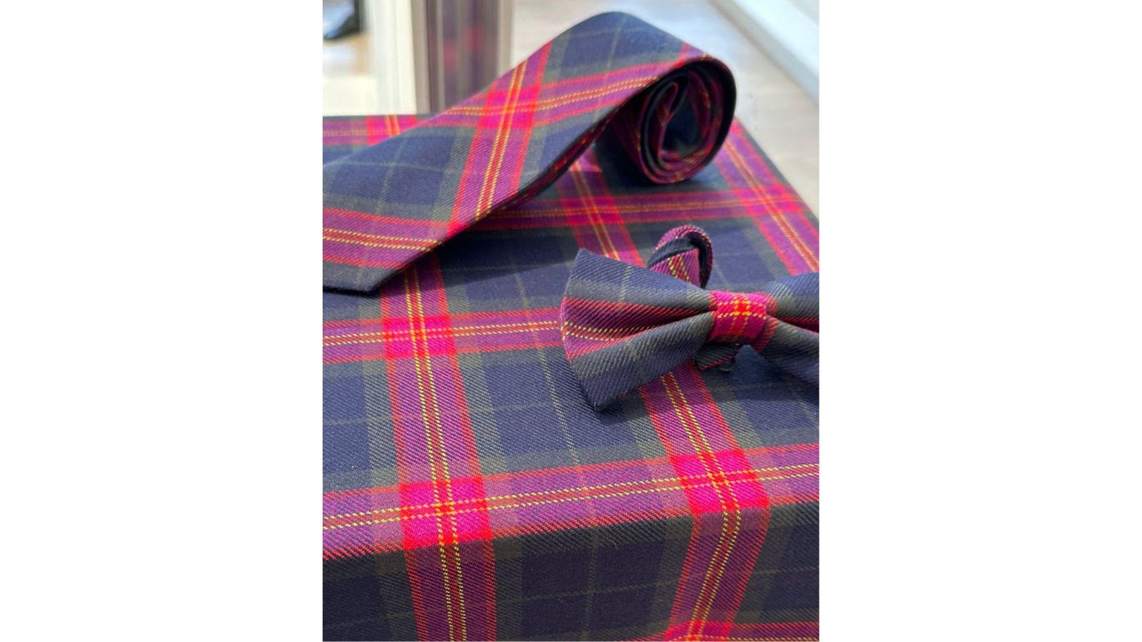 The tartan covers a table, on it are a tie and bow tie made from the tartan. It is dark blue, with threads of red, yellow, green and purple.