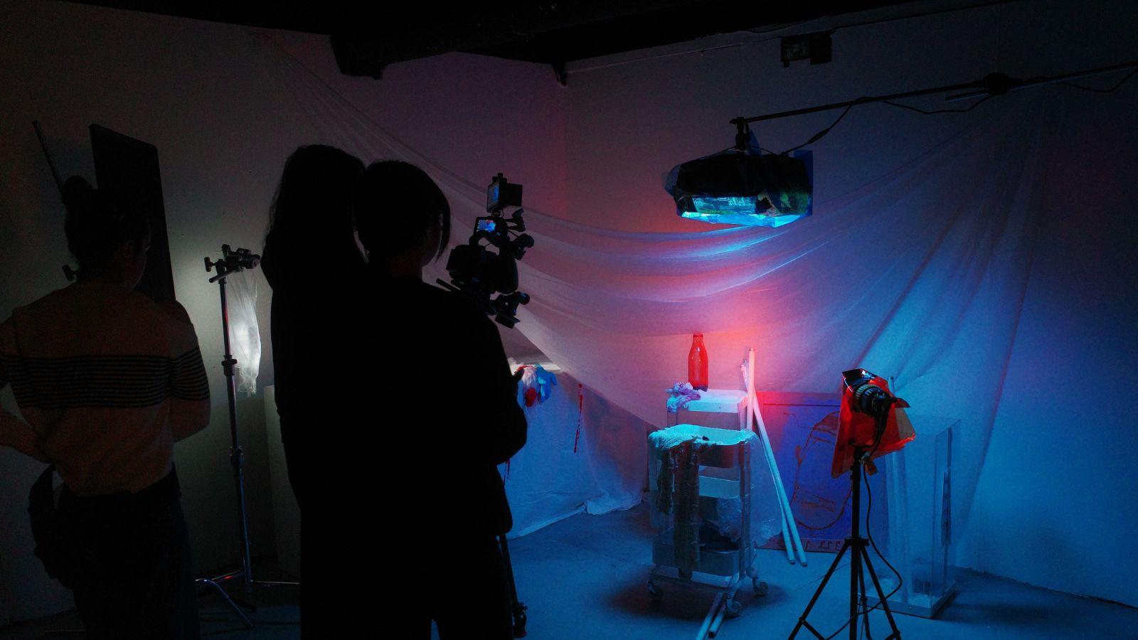 Three students are standing behind a camera, filing a low lit scene which includes a sheet hung up, a set of drawers on wheels and random objects like cloths, bottles, sticks etc littered about.