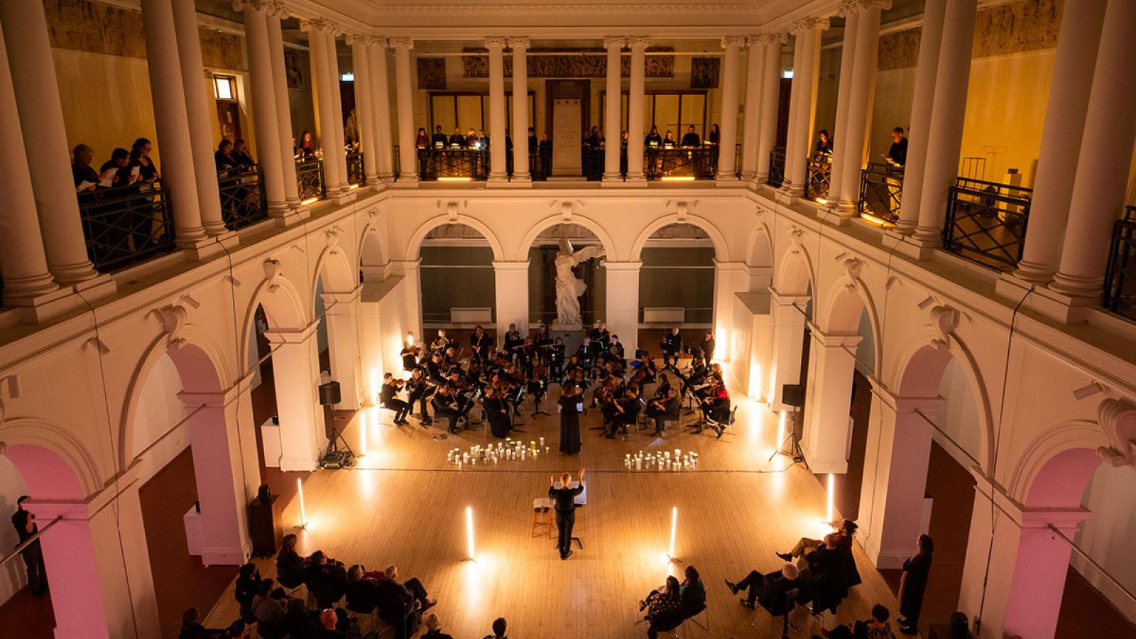 The Sculpture Court, beautifully lit. At one end is an orchestra; standing along the balcony on three sides of the room, is a choir. The conductor's arms are raised.