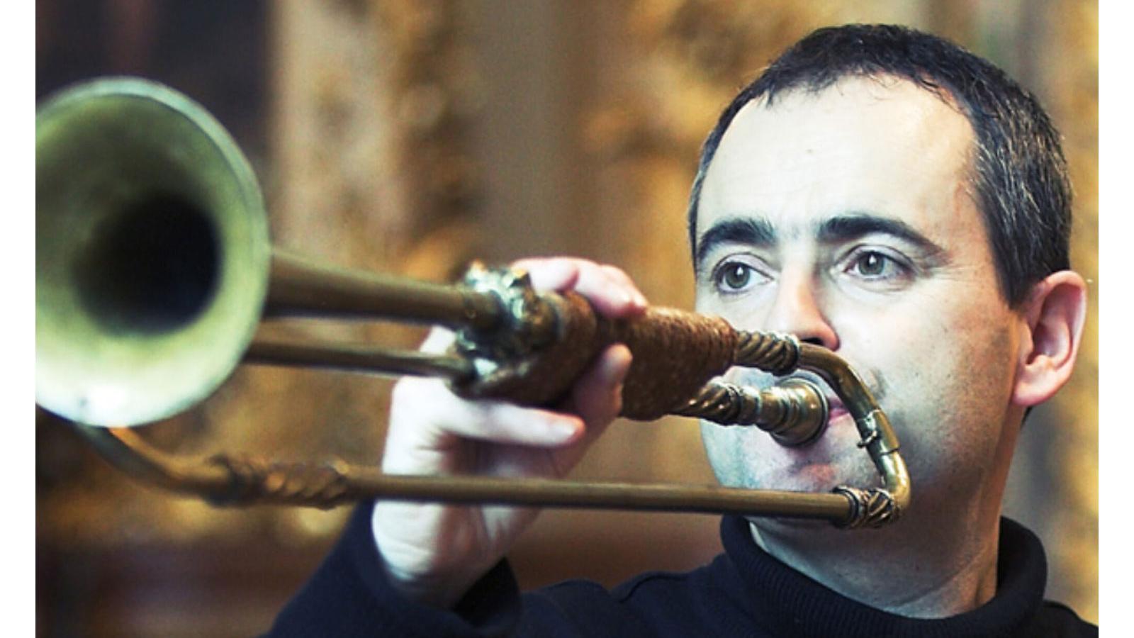 A person with short hair and wearing a dark shirt, blowing into a trombone.
