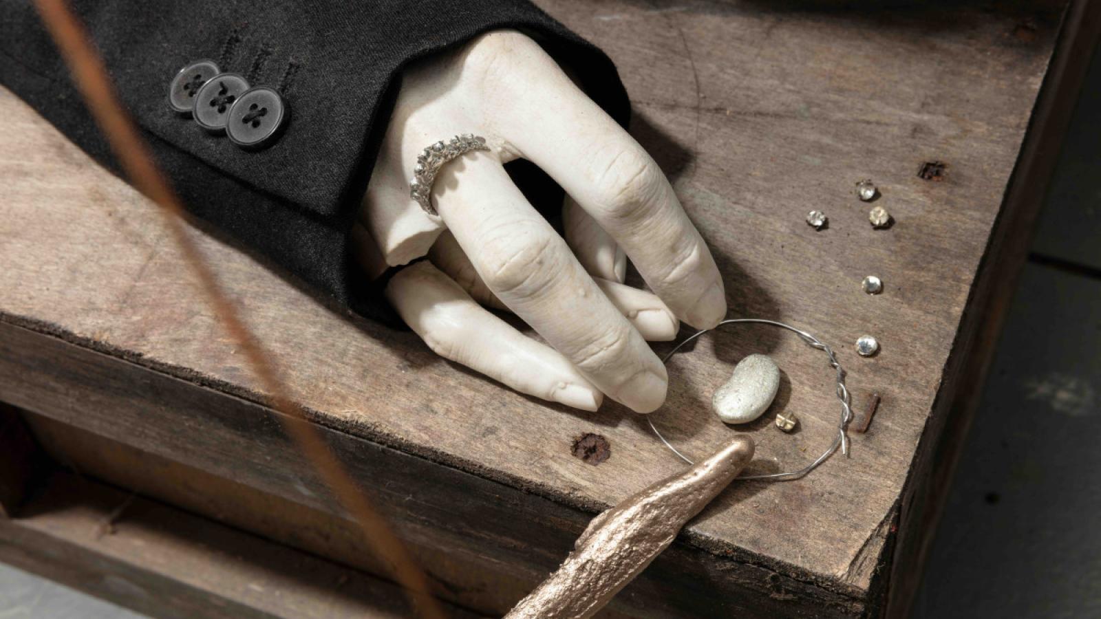 A sculpture of a hand with the third finger broken off. The hand is resting on a piece of wood, with small pieces of metal around it. 