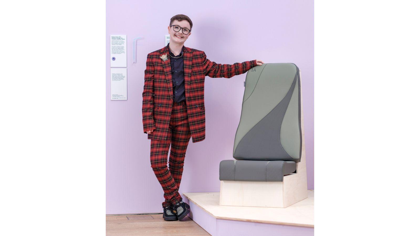 Jamie stands next to his airline seat. He is smiling, and is wearing a red and black tartan suit. The airline seat is a greenish grey, with a design including two curved lines, with a darker colour between them.