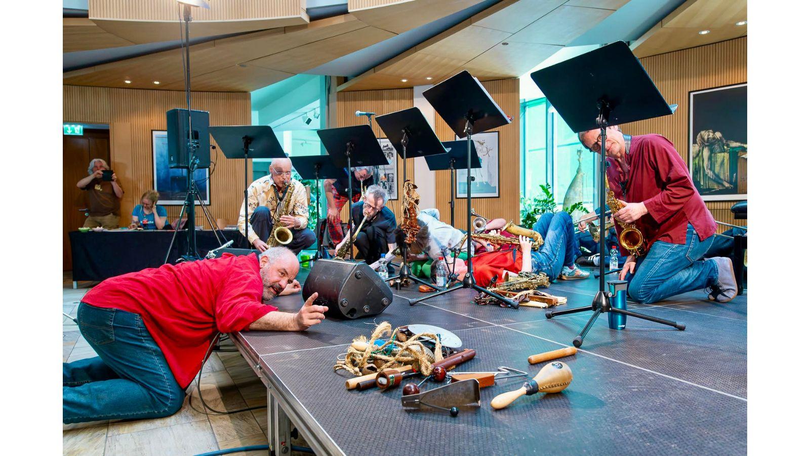 Raymond kneels on the floor in front of the stage, leaning onto the stage. On stage, five saxophonists are playing, all in different positions - kneeling, crouching, lying down. Also on stage are a collection of small instruments, and a row of music stands.