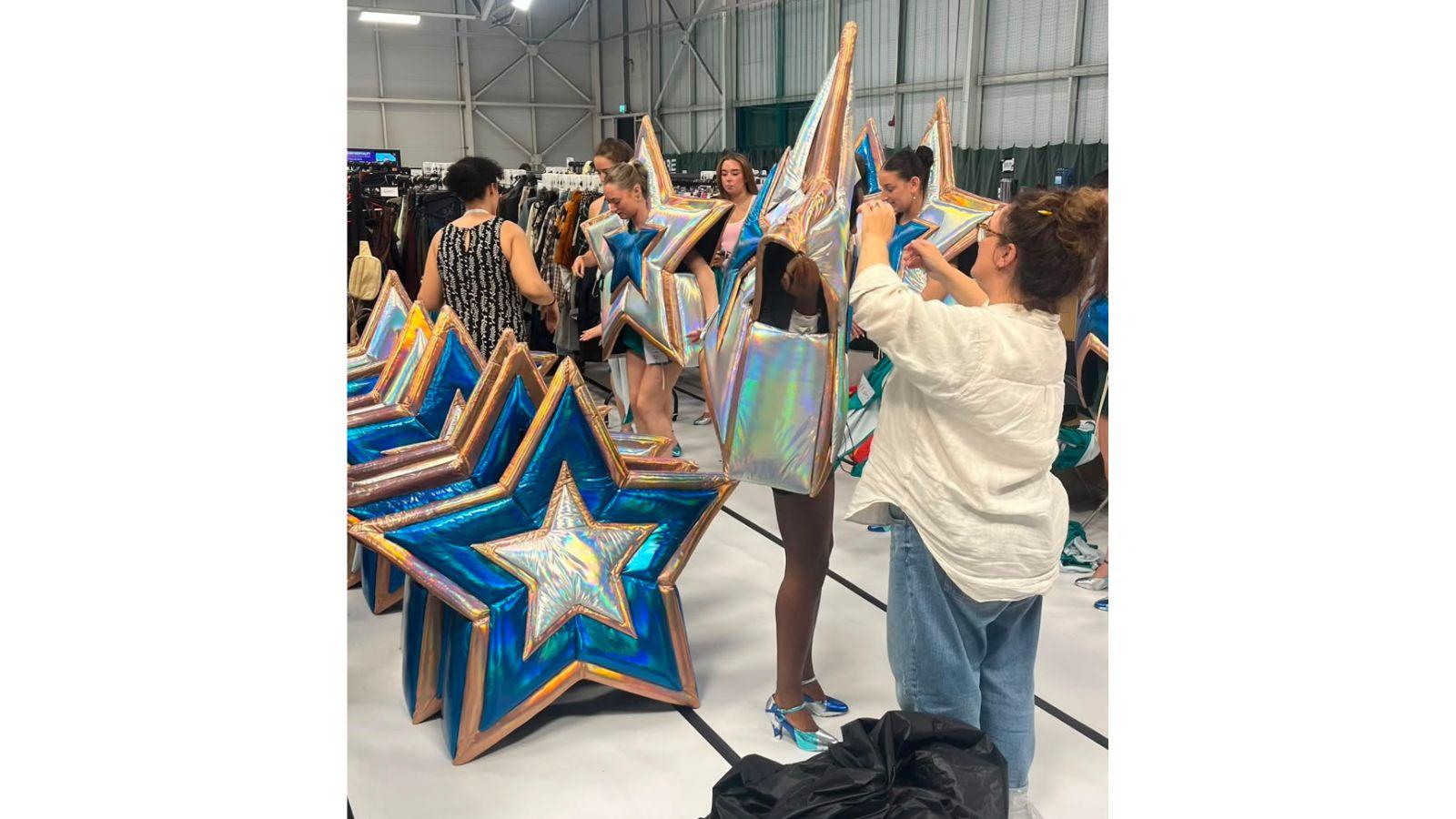 In a large warehouse, costume staff help performers into star-shaped, blue and silver costumes.
