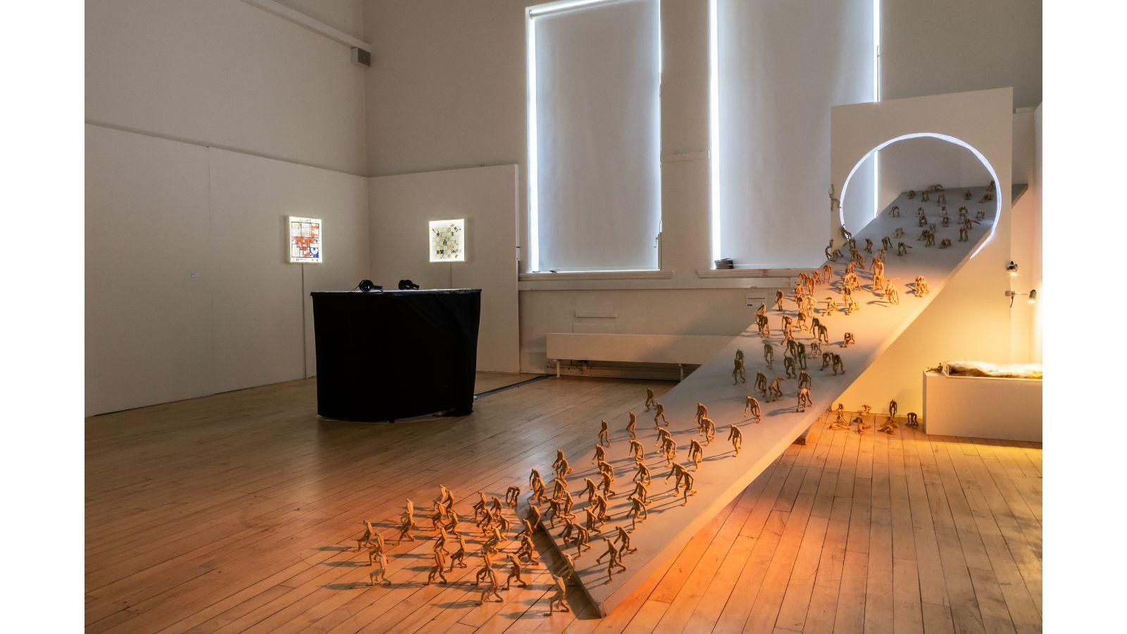 A studio at the Graduate Show, with low lighting. A ramp extends from the floor up to an illuminated circle. Lots of tiny figurines are descending down the ramp.