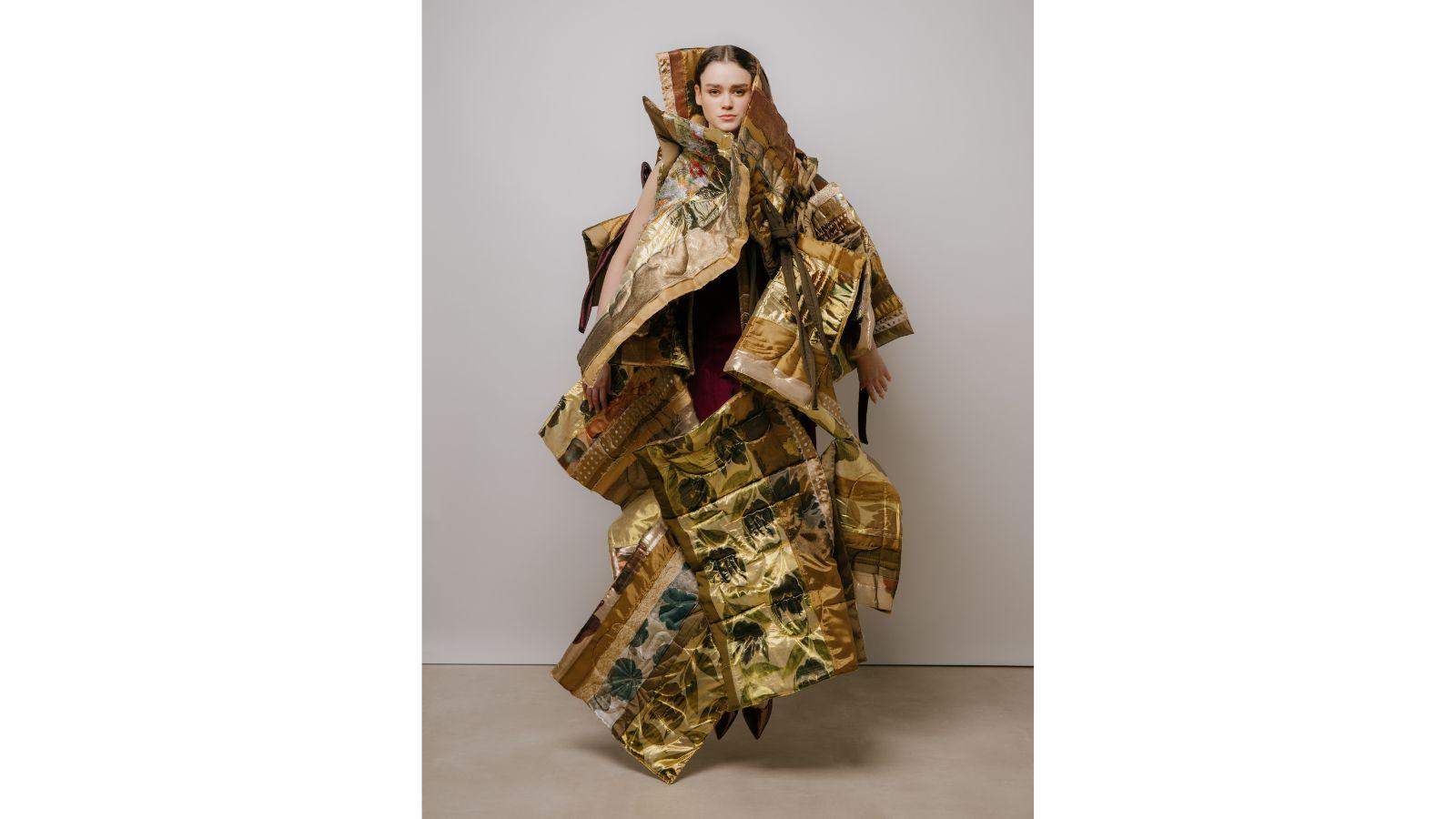 A model wears an outfit made of quilted golden patterned panels, hanging in a plethora of shapes and angles. She is jumping, both toes clear of the ground.