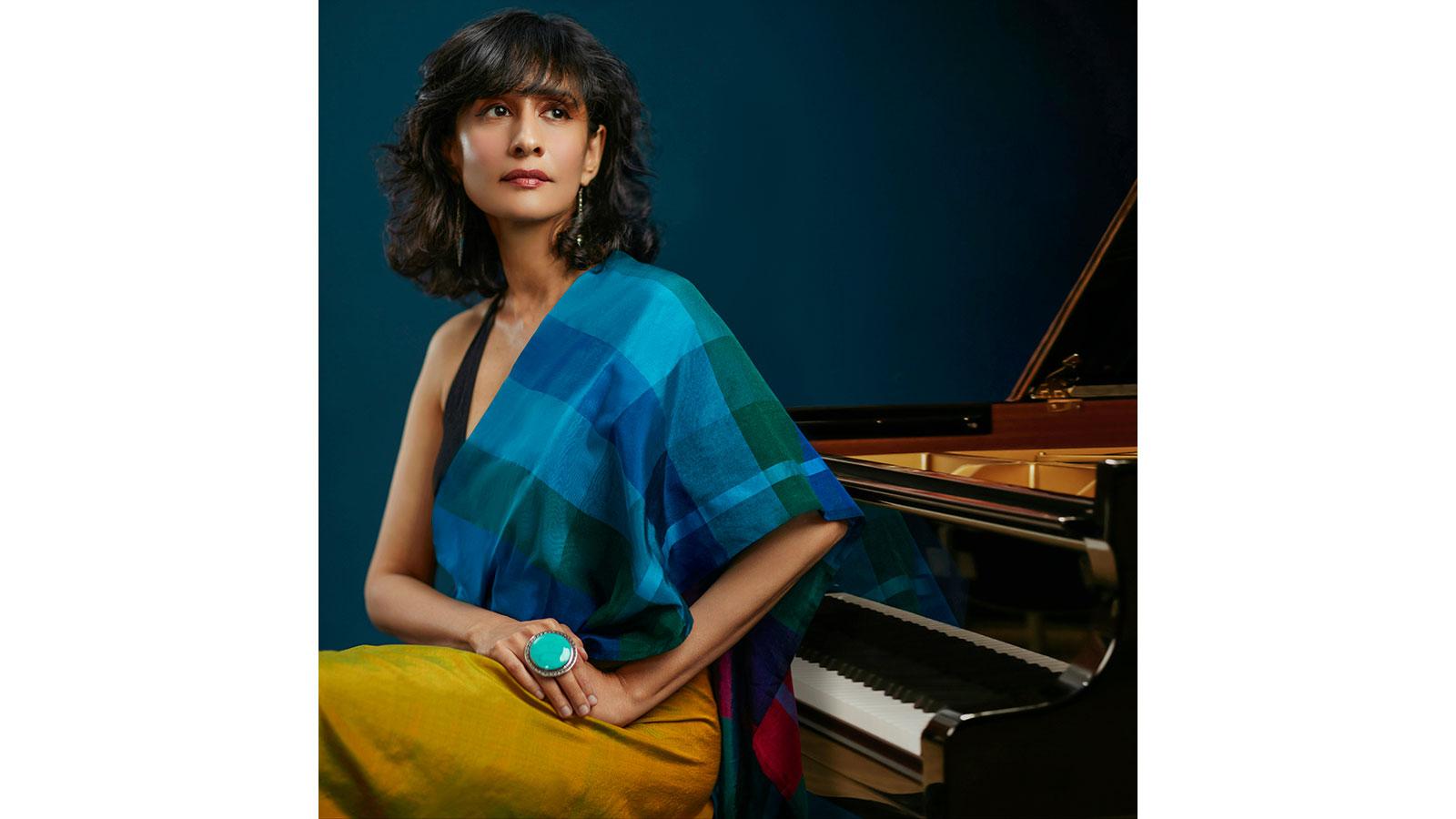 Pianist Dr Tanya Ekanayaka sits by a grand piano. They wear a blue checked shawl and yellow skirt.